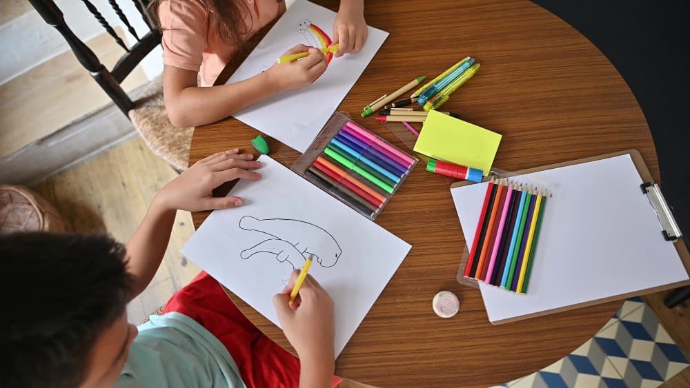 Two children drawing with markers and colored pencils at a kitchen table